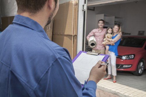 A professional house removals scene in Battersea showing a man in a blue shirt holding a clipboard and pen, preparing to document belongings before a move. In the background, a couple stands outside a residential property, watching the process, with a young girl holding a small dog beside them. The driveway features a red car parked near the entrance, and several cardboard boxes are stacked nearby, likely filled with household items. The house's entrance is partially visible, with an open doorway leading into a hallway or interior room. The lighting suggests daytime with natural light illuminating the scene. This setting highlights the typical environment for local removals services offered by Battersea Man with Van, including careful documentation and attentive service as part of a residential move within the Battersea area or nearby postcodes, emphasizing professionalism and readiness for house relocations.