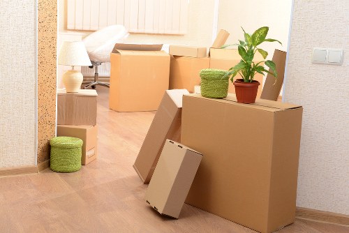 The image depicts a room with several packed cardboard moving boxes of various sizes, some stacked and others placed individually, with visible tape seals and plain brown surfaces. A small green woven storage basket rests on top of a large cardboard box, next to a potted plant with broad green leaves, which sits on another box at waist height. In the background, a portion of a living space is visible, including a white sofa with a beige throw pillow, a small wooden side table with a white lamp, and a light wood floor. The room features neutral wallpaper on the walls, a textured wall panel near the doorway, and a visible light switch. The scene suggests an active house removal or relocation process within a residential property in Battersea, with natural, soft indoor lighting illuminating the scene, emphasizing the arrangement of the boxes and household items prepared for moving. The overall setup aligns with professional house removals services offered by Battersea Man with Van, highlighting typical packing and organizational tasks involved in domestic relocations in the area.