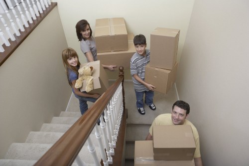 A family of four, including a woman with long dark hair, a young girl with long red hair, a boy with short dark hair, and an adult man, are positioned on a residential staircase landing in a home, preparing for a house move. The woman is holding a cardboard box with a teddy bear inside, while the girl and boy each hold one or two cardboard boxes. The man is standing at the bottom of the staircase, also holding a cardboard box. Behind them are several large cardboard boxes, some sealed and stacked, ready for moving. The staircase features a wooden handrail and white balusters, with neutral-colored carpeted stairs. The under-stair area is part of an interior household space with cream-colored walls, a small window with a wooden ledge on the left, and soft natural lighting. The scene depicts the typical process of packing and relocating belongings from a house in Battersea or nearby London postcodes, with household items such as boxes, boxes, and soft toys being prepared for transport during a professional removals service by Battersea Man with Van.
