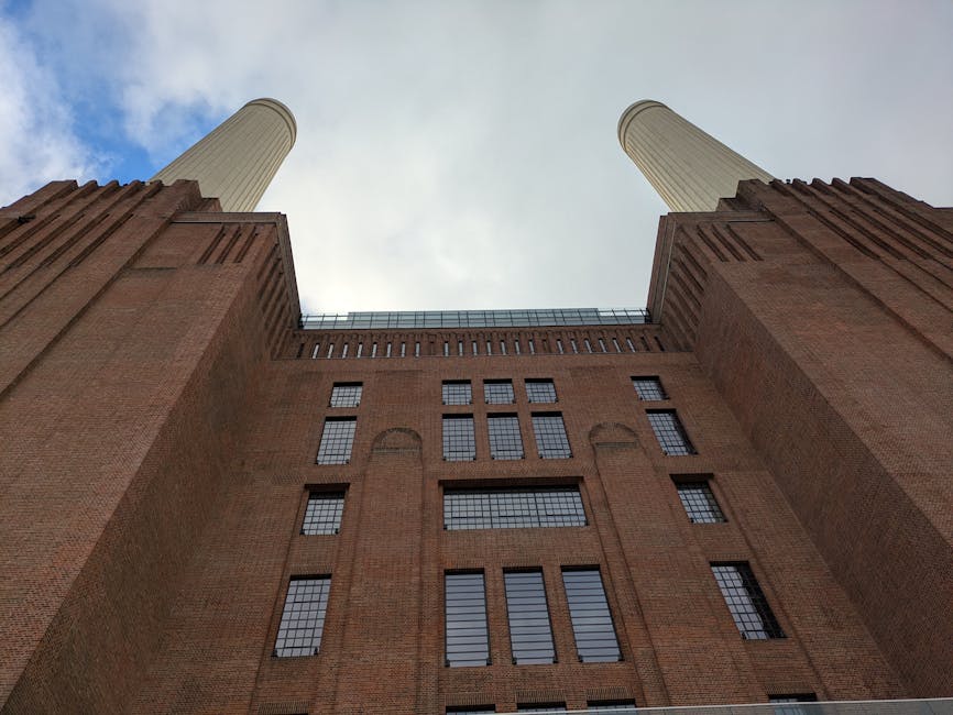 A low-angle view of Battersea Power Station's iconic brick facade with two tall, white chimneys rising against a clear blue sky; the scene depicts the exterior of a historic high-rise building with la